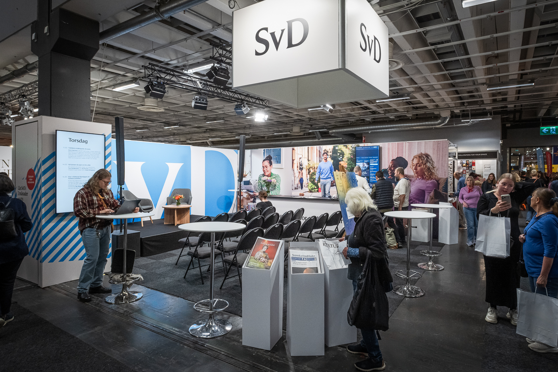 Exhibit booth with SvD signs, chairs, tables with newspapers, and people interacting in a busy indoor event space. Bright lighting and large photos decorate the area.