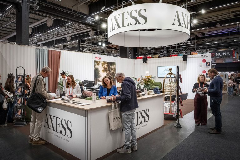People interact at an AXESS magazine booth at an indoor event. The booth features magazines, brochures, and a large circular AXESS sign hanging above. The environment is modern and busy, with several attendees.