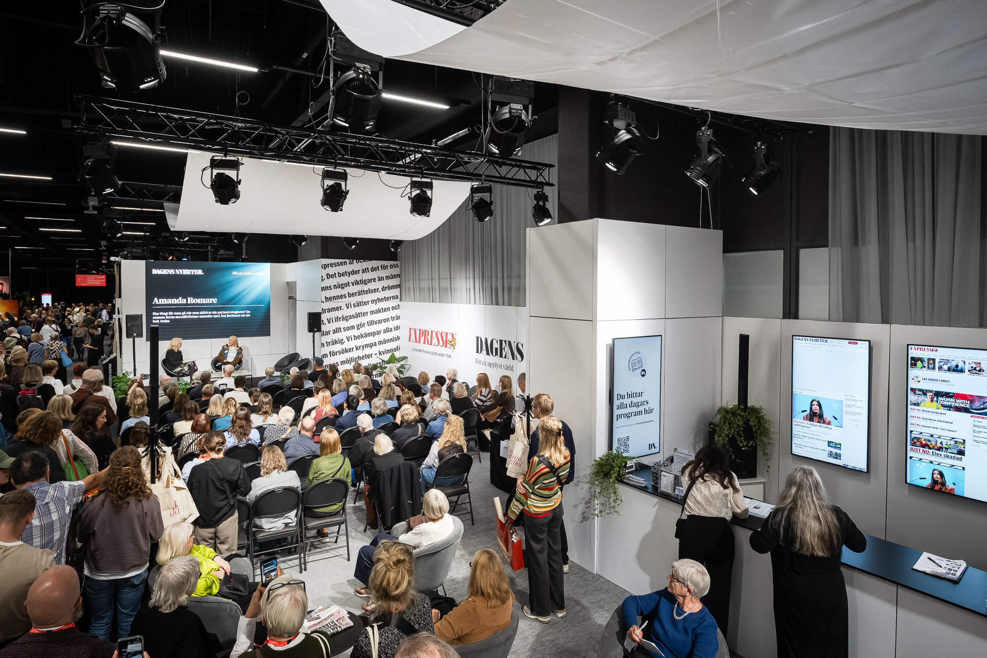 A crowded indoor event with many people seated and watching a speaker on stage. Large screens display information, and some attendees are standing or taking photos. The setting is modern with bright lighting and white decor.