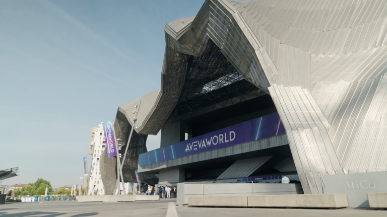 Futuristic building with metallic, angular architecture and AVEAWORLD banners, under a clear blue sky. A few people stand near the entrance.