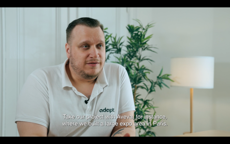 A man in a white adapt polo shirt sits indoors near a plant and lamp, speaking. Subtitles read: Take our project with Aveva for instance, where we built a large expo area in Paris..