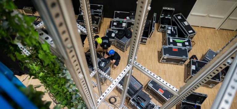 Two workers in helmets are surrounded by equipment cases, viewed from above through metal beams, preparing or organizing gear on a wooden floor. Green plants hang on the side.