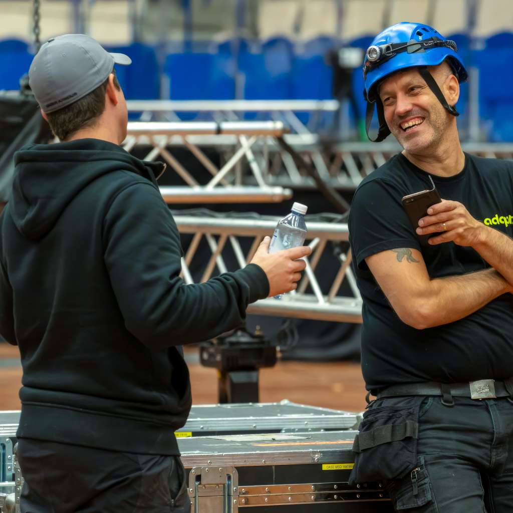 Two men in work clothes chat and smile near metal trusses. One holds a phone and wears a blue safety helmet, while the other holds a water bottle. They appear relaxed in an indoor industrial setting.