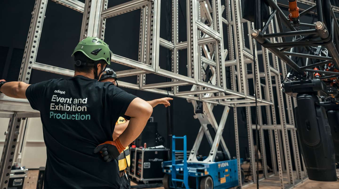 Two workers wearing helmets stand near a large metal framework. One points towards the structure while the other listens. The back of one shirt reads Event and Exhibition Production. Industrial equipment is visible nearby.