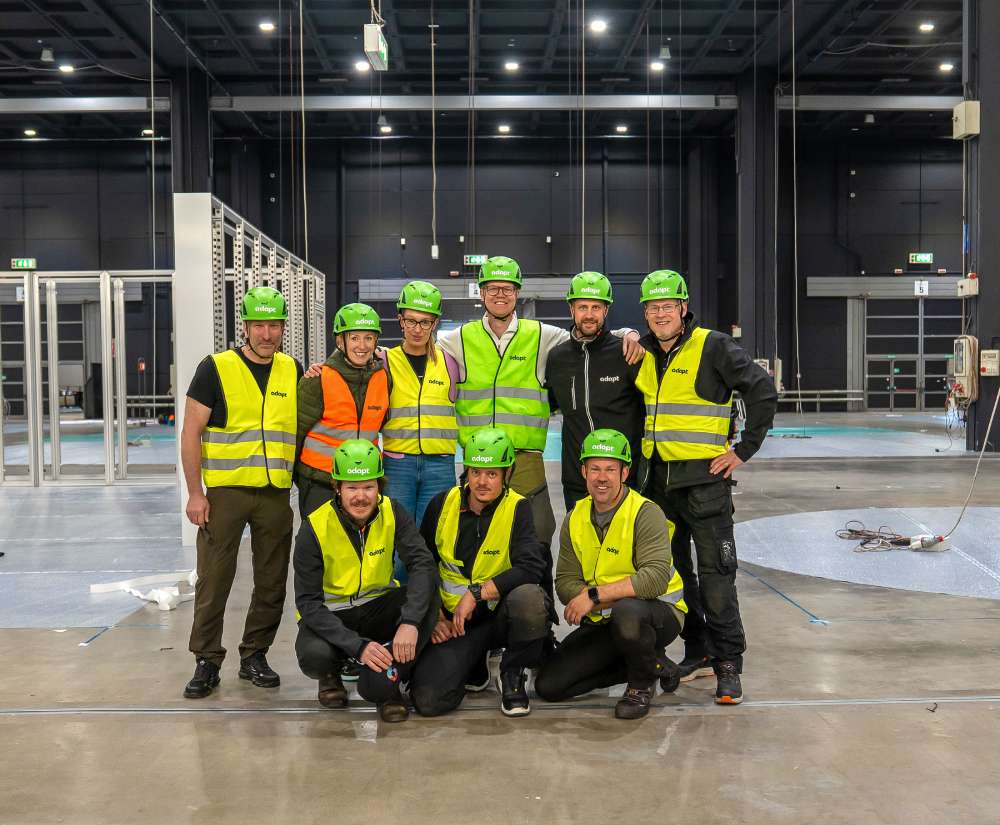 Nine people in safety vests and green helmets pose inside an industrial building under construction. Some are standing, others kneeling, with unfinished metal structures and tools visible in the background.