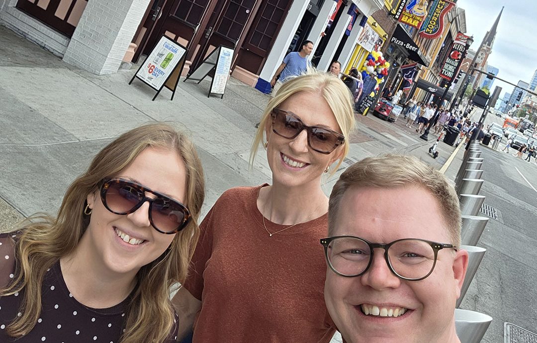 Three people pose for a selfie on a sidewalk in front of colorful neon signs and storefronts on Broadway in Nashville.