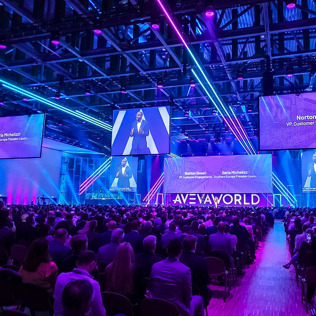 A large audience sits in a dimly lit conference hall watching a keynote presentation on multiple overhead screens displaying speakers and the text AVEVAWORLD.