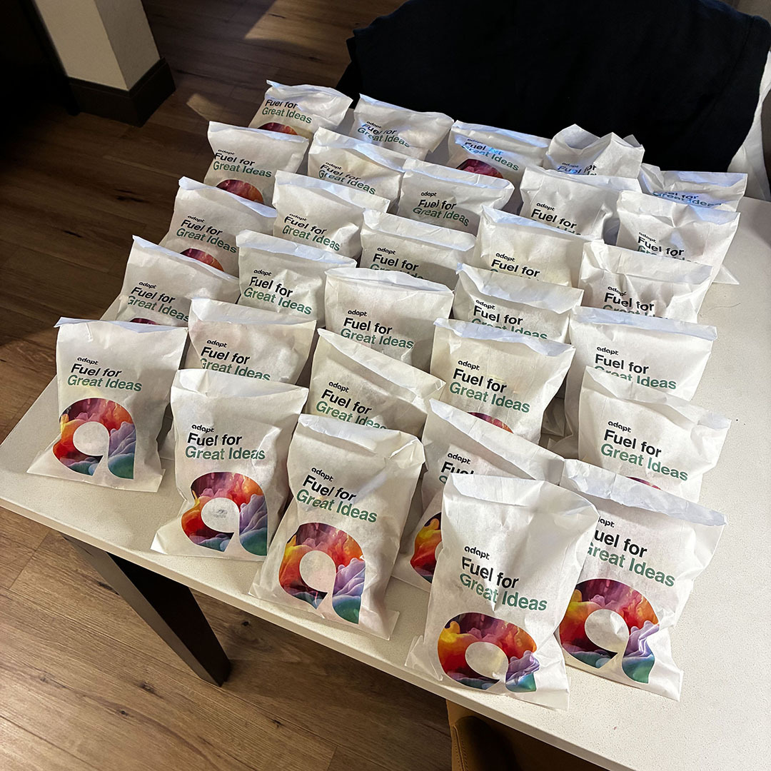 A tabletop covered in rows of white paper gift bags, each printed with the text adopt Fuel for Great Ideas and a colorful abstract graphic.
