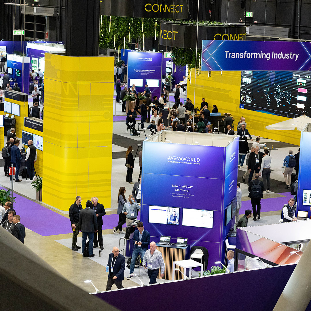 People walk through a large, multi-level trade show floor featuring bright yellow pillars and blue booths branded with AVEVA WORLD and Transforming Industry signage.