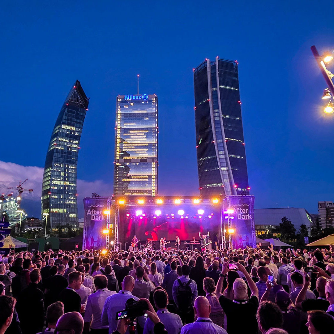 A large crowd watches a live musical performance on an outdoor stage at night, set against a backdrop of illuminated skyscrapers.