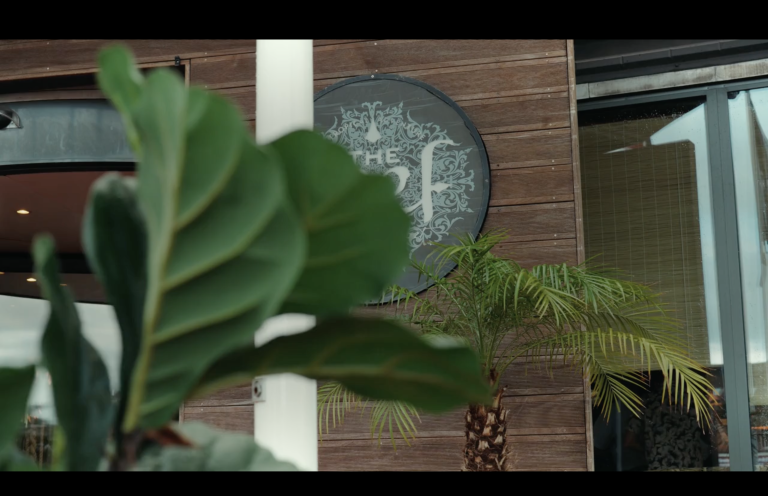 A circular sign on a wood-paneled wall that reads The Reef, partially obscured by a foreground fiddle-leaf fig plant and a smaller palm tree.