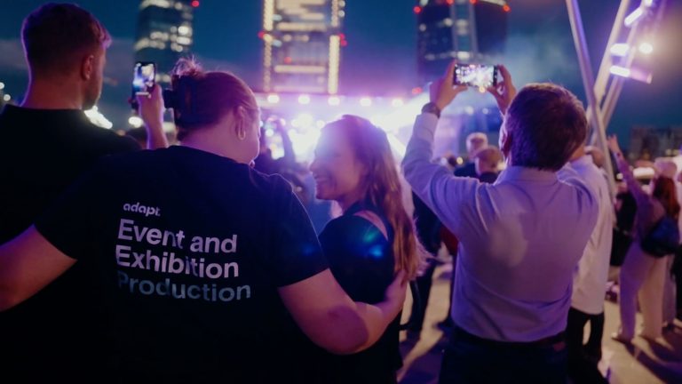 A crowd of people at an outdoor evening event, with one woman wearing a shirt that says adapt. Event and Exhibition Production.